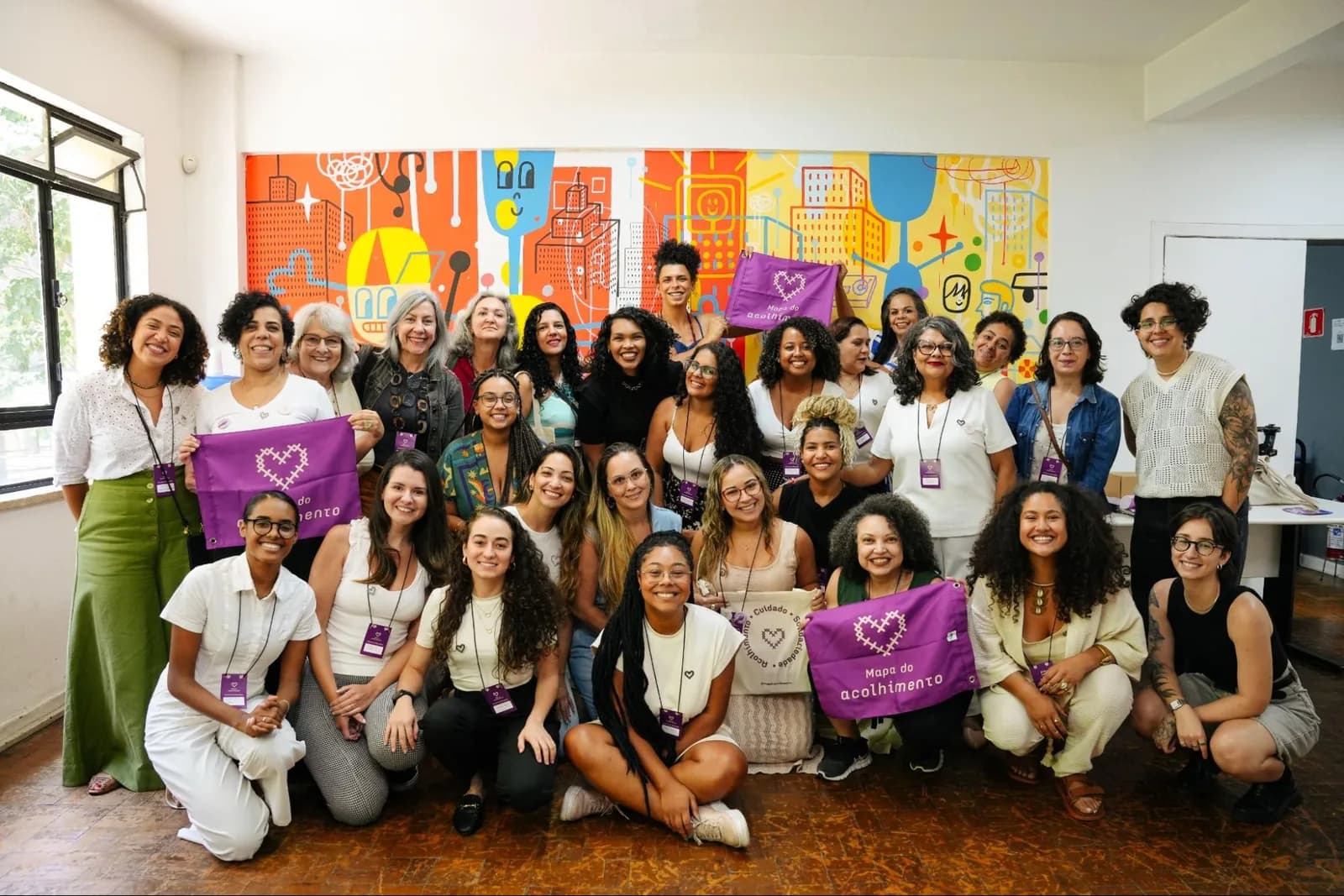 Around thirty Mapa volunteers and team members, many holding purple Mapa do Acolhimento flags, smiling for a group photo in front of a colourful mural.