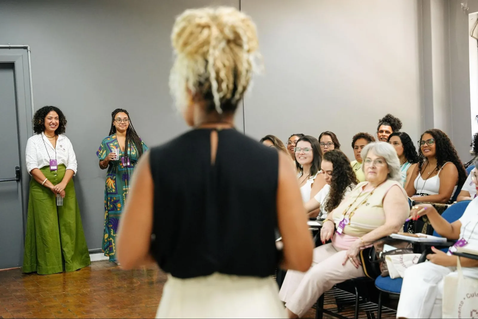 A facilitator, seen from behind, addresses a room of seated women at a Mapa event while others look on and smile.